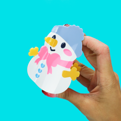 A hand holding a small snowman-shaped item against a bright turquoise background. The snowman is white with a pink scarf, yellow hands and beak, black eyes, and a light blue hat.