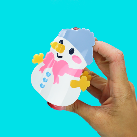 A hand holding a small snowman-shaped item against a bright turquoise background. The snowman is white with a pink scarf, yellow hands and beak, black eyes, and a light blue hat.