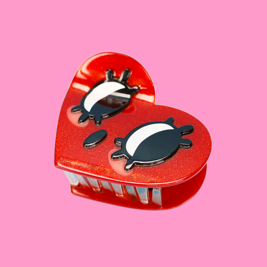 A hand holding a red glittery heart-shaped hair claw clip with black and white cartoon eyes, set against a solid pink background.