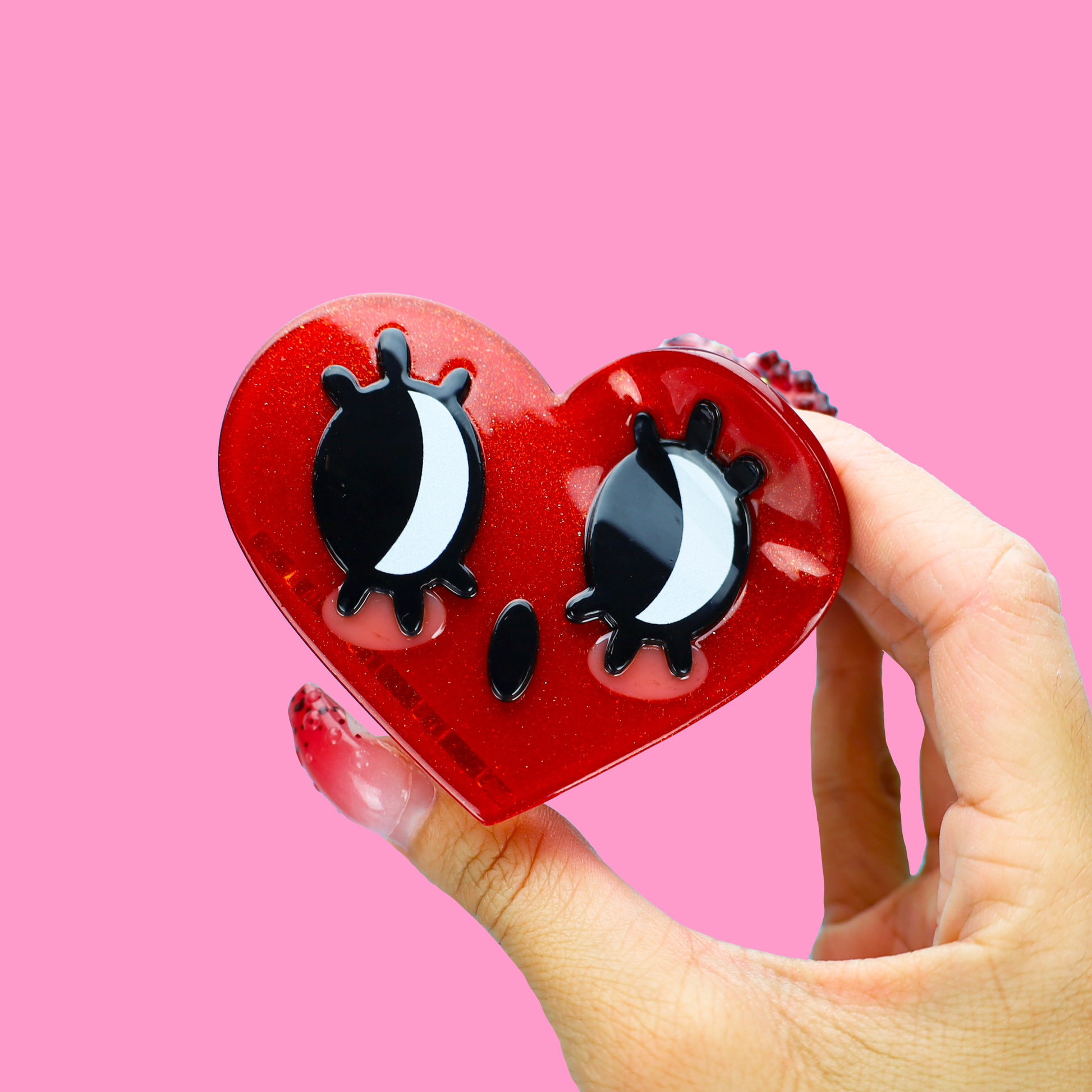 A hand holding a red glittery heart-shaped hair claw clip with black and white cartoon eyes, set against a solid pink background.