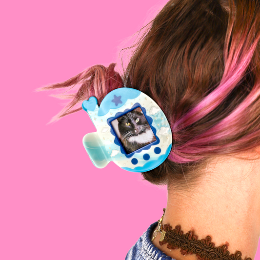 A close-up lifestyle shot of a person's hair secured with a light blue and white tamagotchi-shaped hair clip, set against a solid pink background.