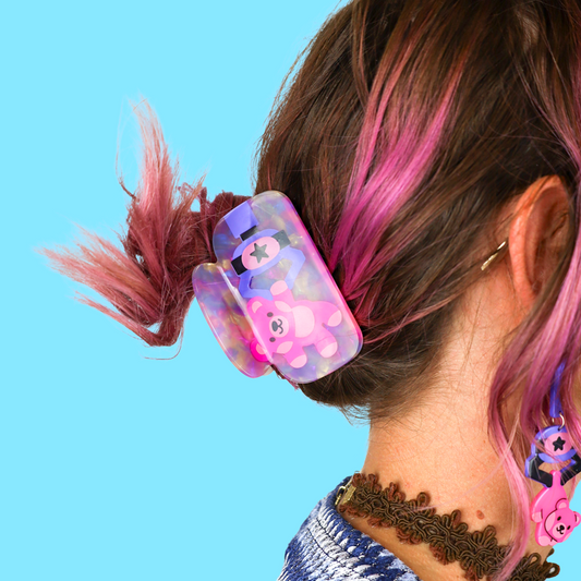 A close-up lifestyle shot of a person's hair secured with a translucent purple and pink bear-print hair claw clip, set against a solid light blue background.