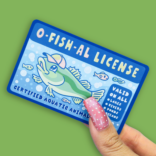 A hand with pink glitter nail polish holds an O-FISH-AL license card featuring a cartoon green fish wearing a cap on a light blue water-themed background, set against a solid green backdrop.