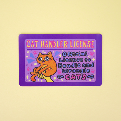 A rectangular novelty card or sticker with a purple border and a cartoon orange cat, reading 'CAT HANDLER LICENSE' and 'Official License to Handle and Wrangle CATS,' shown on a pale yellow background.