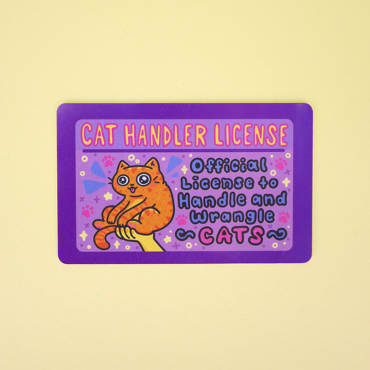 A rectangular novelty card or sticker with a purple border and a cartoon orange cat, reading 'CAT HANDLER LICENSE' and 'Official License to Handle and Wrangle CATS,' shown on a pale yellow background.