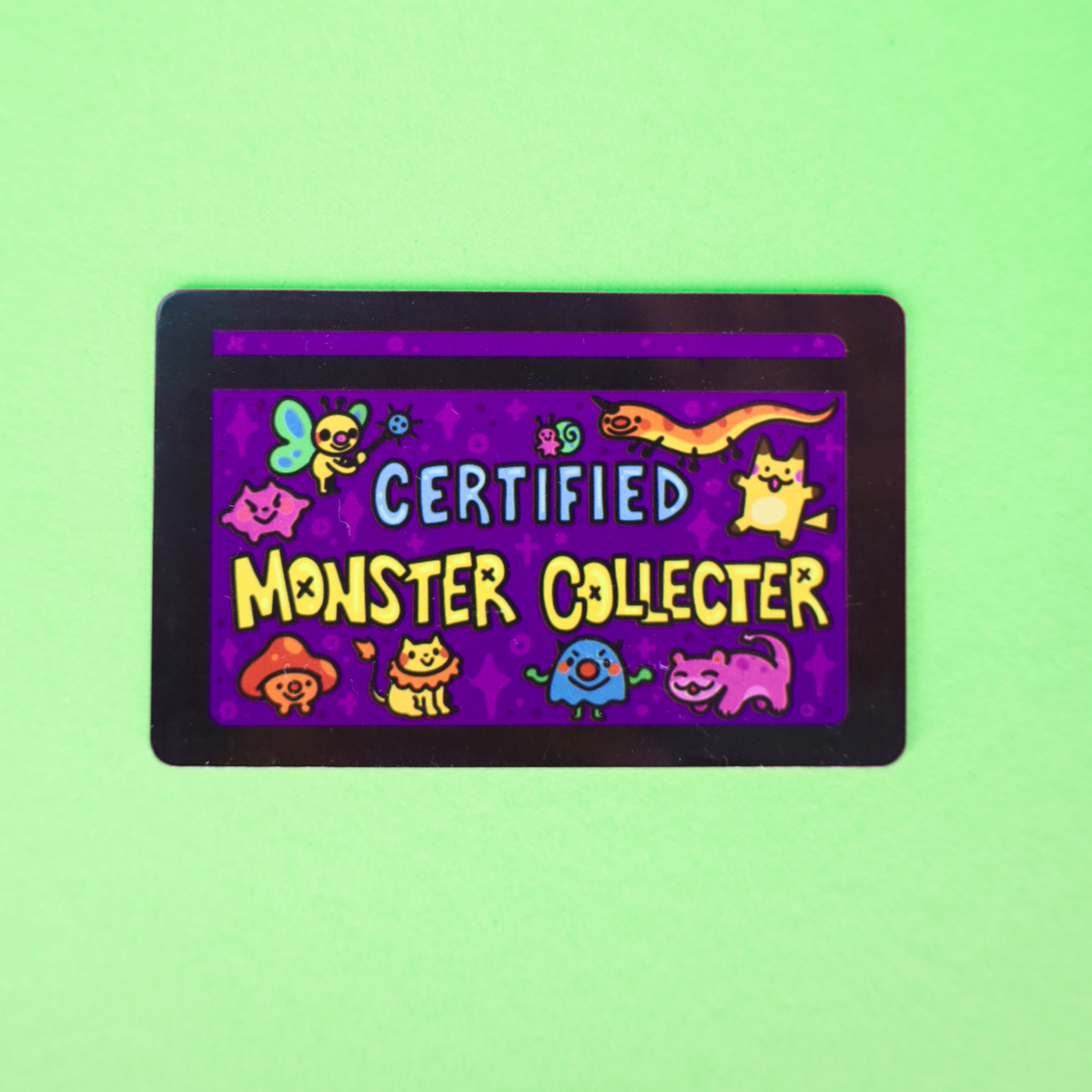A rectangular collectible card with rounded corners featuring a purple background and cartoon monster illustrations, with the text 'CERTIFIED MONSTER COLLECTOR' in the center, shown on a light green background.
