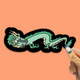 A die-cut sticker featuring a stylized green dragon with cream details and a black outline, held by a hand against a solid peach background.