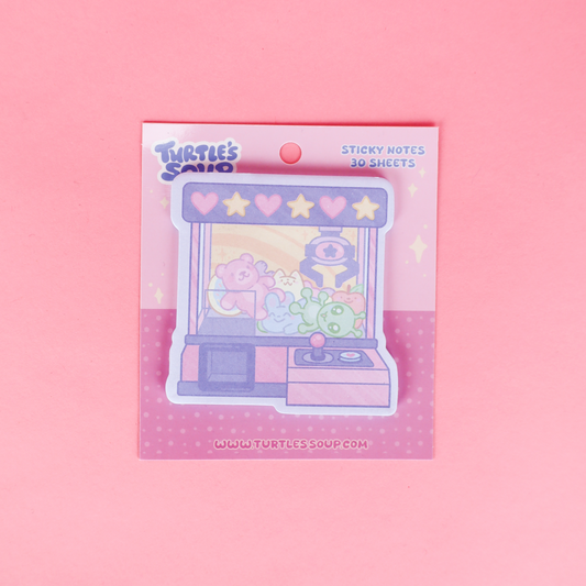 A package of 'Turtle's Said' sticky notes featuring a pastel claw machine illustration, styled in a flat-lay on a pink background with a pink marker, pencil, purple pen, and eraser.