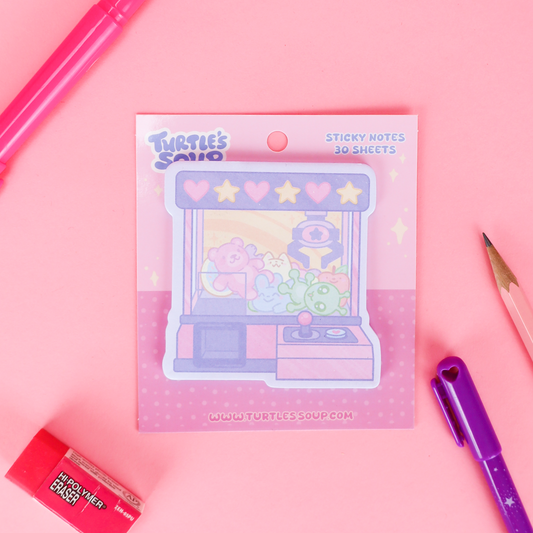 A package of 'Turtle's Said' sticky notes featuring a pastel claw machine illustration, styled in a flat-lay on a pink background with a pink marker, pencil, purple pen, and eraser.