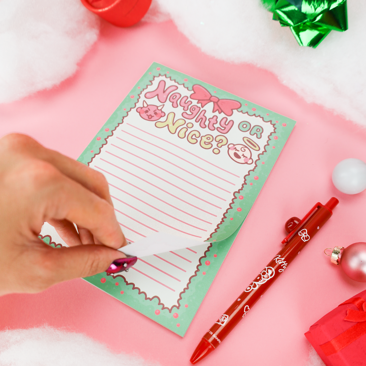 A hand peels back a transparent protective cover from a pastel green and pink notepad that reads 'Naughty or Nice?'. A red decorative pen and holiday ornaments are placed nearby on a pink surface with white faux snow.