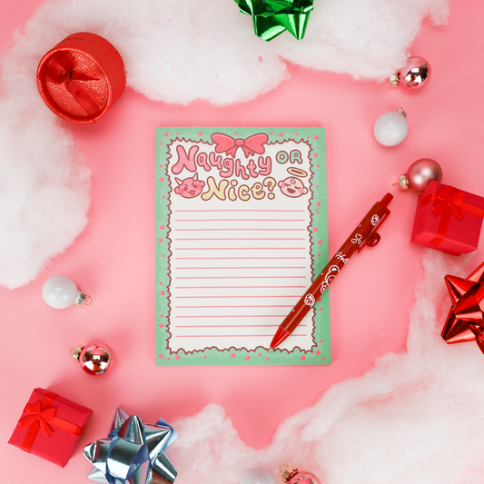 A festive flat-lay of a lined notepad with the words 'Naughty or Nice?' in pink lettering, surrounded by Christmas decorations including a red pen, ornaments, gift boxes, and faux snow on a pink background.