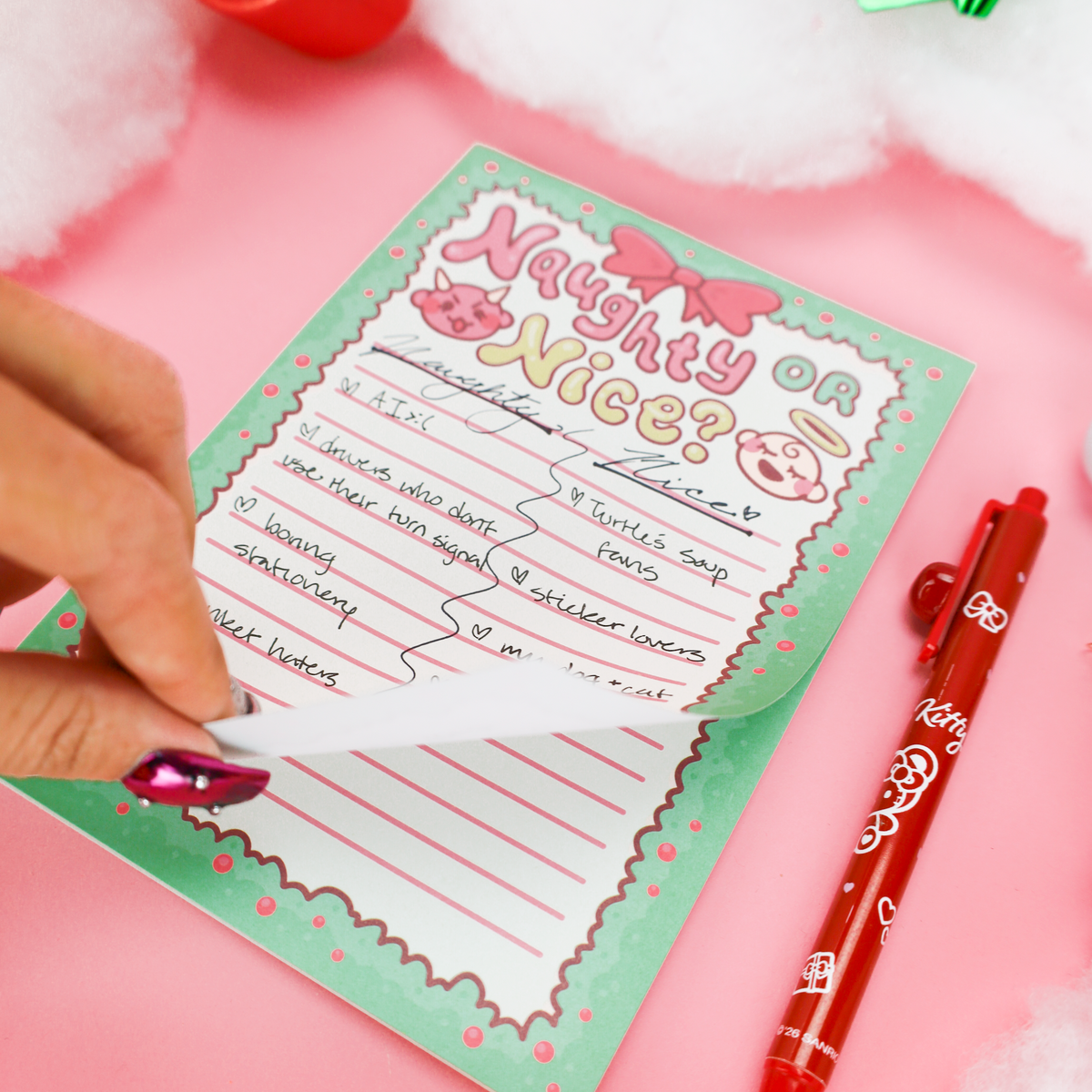 A hand holds a transparent peel-away sticker sheet from a 'Naughty or Nice?' notepad with pink and green borders, placed on a pink background next to a red pen.
