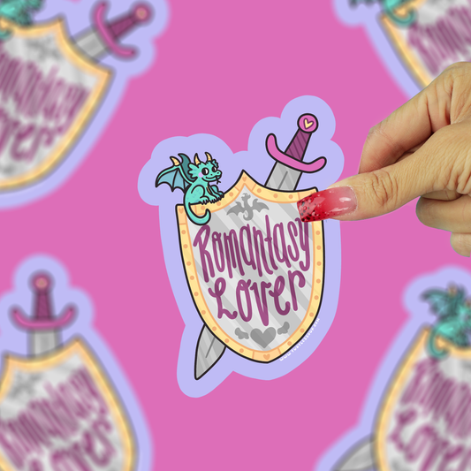 A hand with red nail polish holds a die-cut sticker featuring a fantasy shield with the words 'Romantasy Lover' in purple script, a silver sword, and a small green dragon, against a pink background with blurred repeating versions of the same sticker.