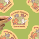 cats surrounding a table playing mahjong that says i love playing meowjong with my friends vinyl sticker being held against a green background