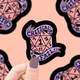 Sticker with a D20 die design and 'Critical Fail' text held by a hand against a patterned background.
