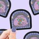 Sticker with a dragon illustration and 'Dice Hoarder' text held by a hand against a purple background with more stickers.