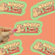 Hand holding a 'Hangry!' sticker with a cartoon dog and food items on an orange background.