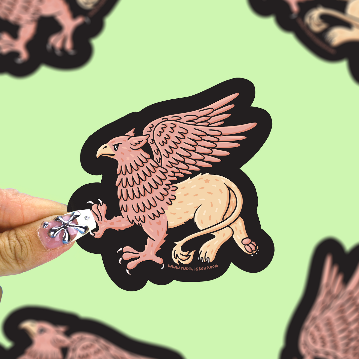 A hand with a glittery nail design holds a die-cut sticker of a griffin against a light green background with other blurred stickers.