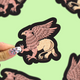 A hand with a glittery nail design holds a die-cut sticker of a griffin against a light green background with other blurred stickers.