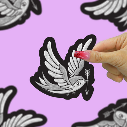A hand with pink glittery nails holds a die-cut sticker of a white and gray dove holding an arrow with a black outline, set against a light purple background with other blurred stickers.