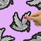 A hand with pink glittery nails holds a die-cut sticker of a white and gray dove holding an arrow with a black outline, set against a light purple background with other blurred stickers.
