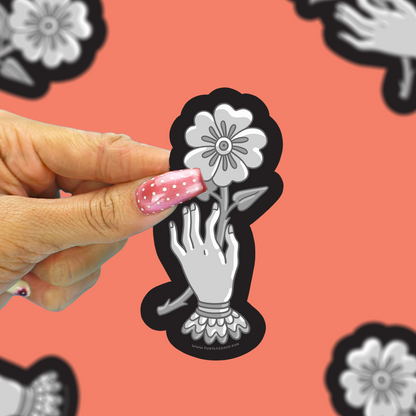 A hand with pink glitter nail polish holds a die-cut sticker featuring a black and white illustration of a hand reaching for a flower. The background is a coral pink with repeated blurred versions of the same sticker design.