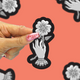 A hand with pink glitter nail polish holds a die-cut sticker featuring a black and white illustration of a hand reaching for a flower. The background is a coral pink with repeated blurred versions of the same sticker design.