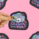 A hand with patterned nail polish holds a black die-cut sticker featuring a skull and books with the text 'Death by TBR' in purple and pink, against a pink background with other copies of the sticker.