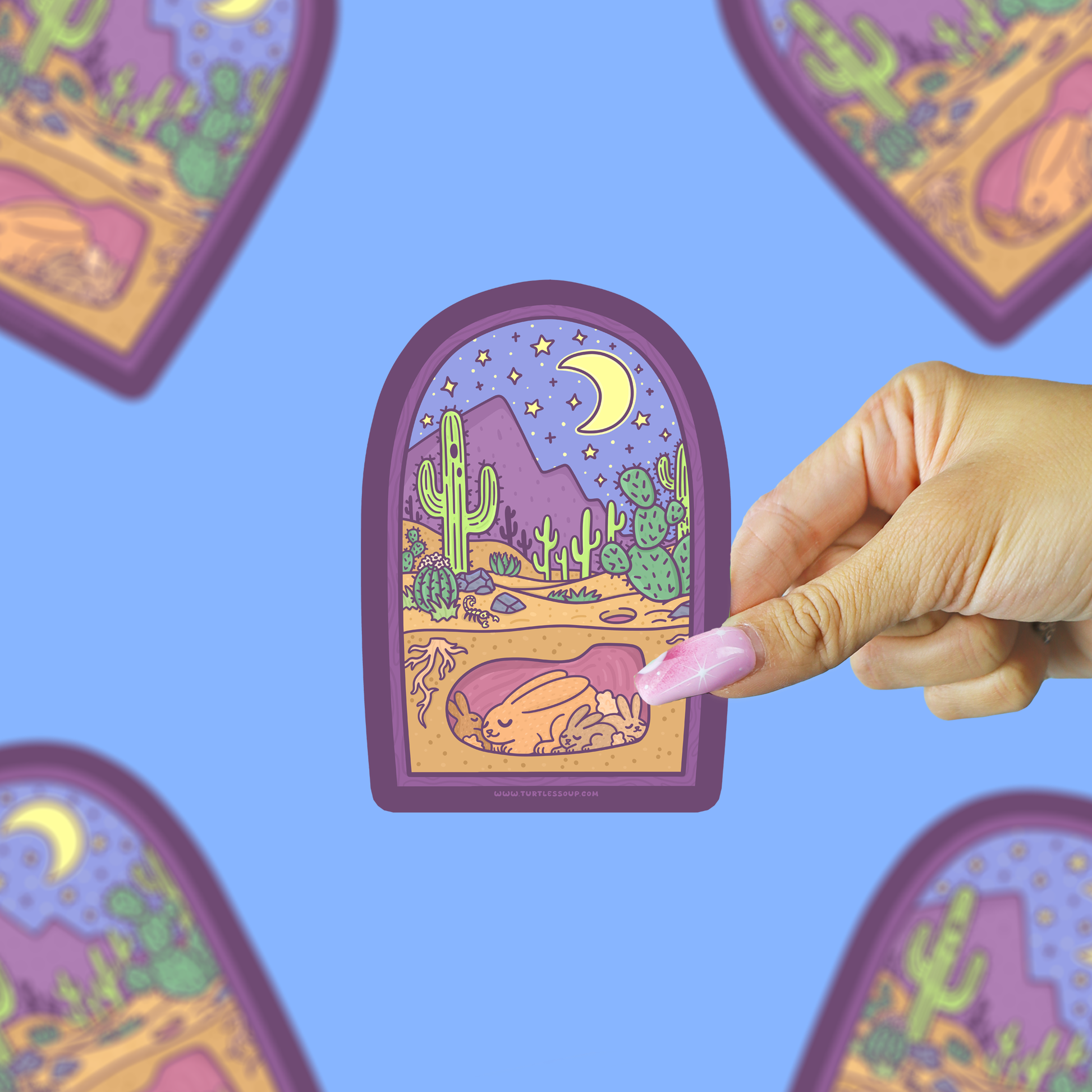 A hand with pink nails holds a die-cut sticker featuring a stylized desert night scene with cacti, mountains, a crescent moon, and a sleeping figure. The background is a solid light blue with blurred duplicate views of the sticker.