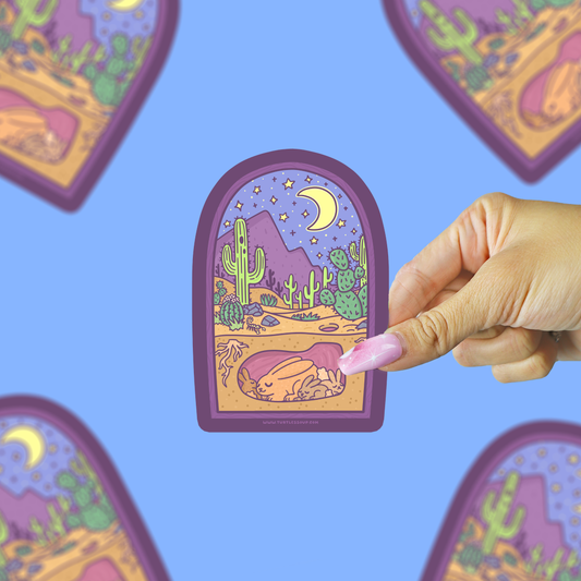A hand with pink nails holds a die-cut sticker featuring a stylized desert night scene with cacti, mountains, a crescent moon, and a sleeping figure. The background is a solid light blue with blurred duplicate views of the sticker.