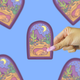 A hand with pink nails holds a die-cut sticker featuring a stylized desert night scene with cacti, mountains, a crescent moon, and a sleeping figure. The background is a solid light blue with blurred duplicate views of the sticker.