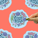 A hand with gold nail polish holds a die-cut sticker that reads 'Friend to Birds' surrounded by colorful illustrated birds on a light blue cloud-shaped background. The sticker is centered against a coral-pink background with partial duplicate sticker images around the edges.