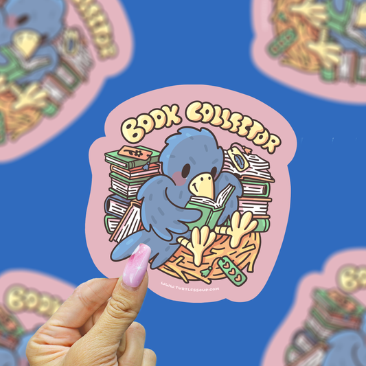A hand with pink manicured nails holds a die-cut sticker featuring a blue bird reading books, with the text 'BOOK COLLECTOR' in yellow. The background is a blue surface with blurred, repeating versions of the sticker.
