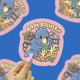 A hand with pink manicured nails holds a die-cut sticker featuring a blue bird reading books, with the text 'BOOK COLLECTOR' in yellow. The background is a blue surface with blurred, repeating versions of the sticker.