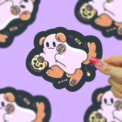 A hand with pink manicured nails holds a die-cut sticker featuring a cute cartoon ghost dog character. The sticker has a black outline, a pale pink body, brown paws and ears, and small gold Halloween-themed details, set against a light purple background with blurred duplicate sticker images.