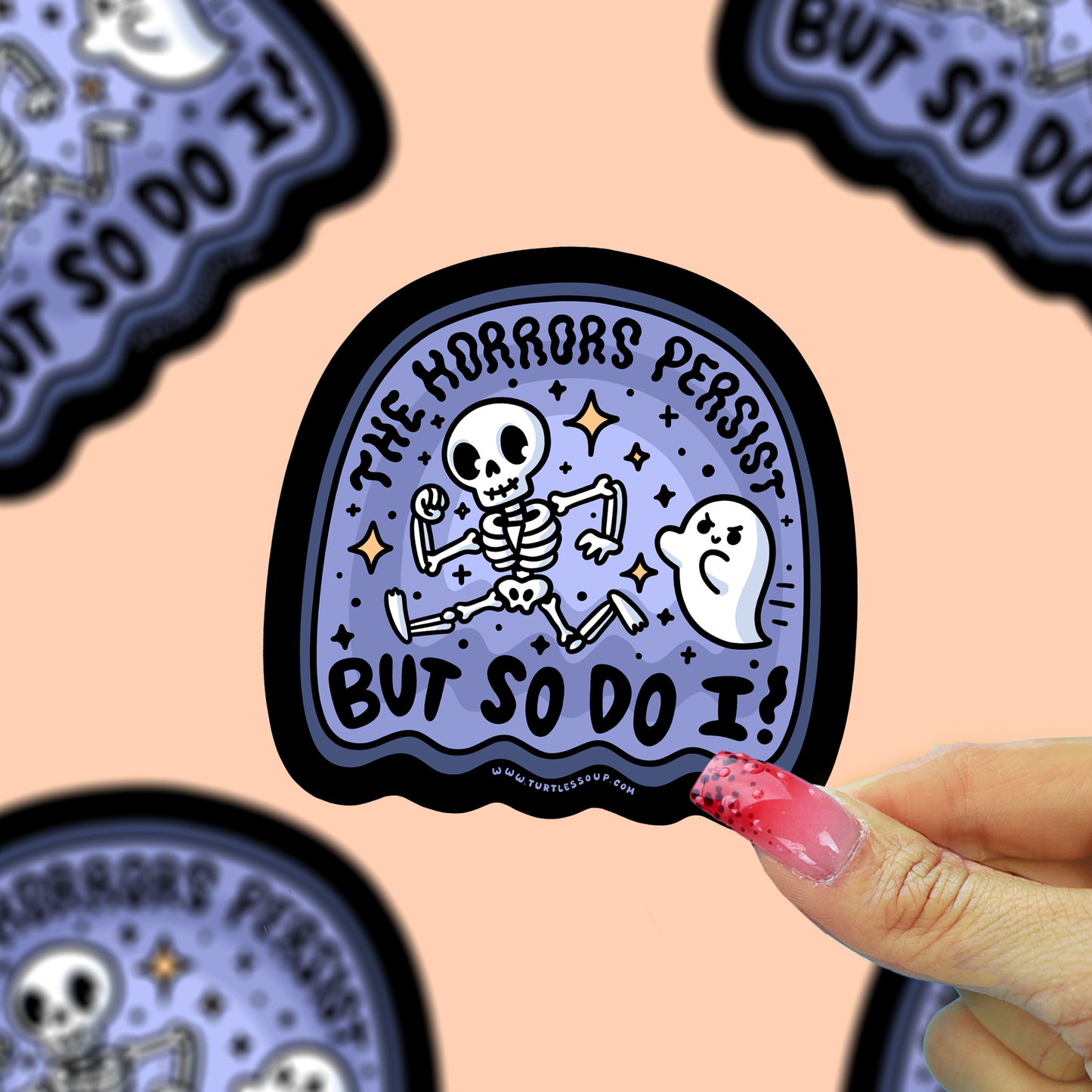 A hand with pink glittery nails holds a die-cut sticker featuring a cartoon skeleton and ghost with the text 'THE HORRORS PERSIST BUT SO DO I!' on a lavender background with a black border. Other similar stickers are partially visible in the background against a peach-colored backdrop.