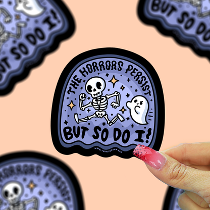A hand with pink glittery nails holds a die-cut sticker featuring a cartoon skeleton and ghost with the text 'THE HORRORS PERSIST BUT SO DO I!' on a lavender background with a black border. Other similar stickers are partially visible in the background against a peach-colored backdrop.