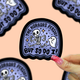 A hand with pink glittery nails holds a die-cut sticker featuring a cartoon skeleton and ghost with the text 'THE HORRORS PERSIST BUT SO DO I!' on a lavender background with a black border. Other similar stickers are partially visible in the background against a peach-colored backdrop.