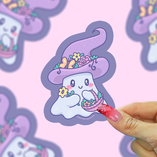 A cute cartoon ghost character sticker with a purple witch hat and flowers, held by a hand with pink glittery nails against a pink background with blurred duplicate stickers.