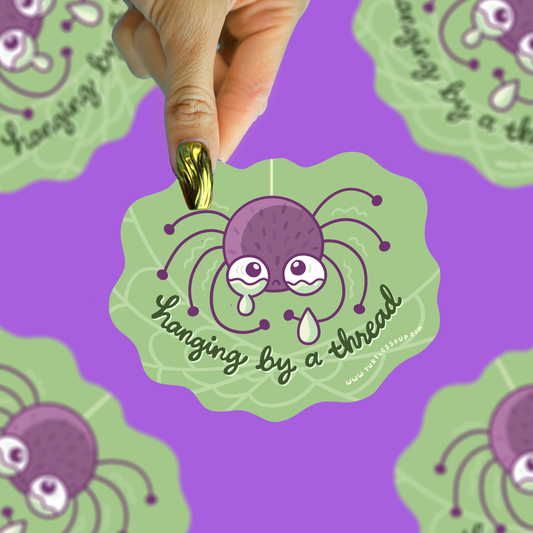 A hand with gold nail polish holds a die-cut sticker featuring a cartoon purple spider hanging by a thread on a light green leaf background. The sticker is shown against a purple background with a repeating pattern of the same design.