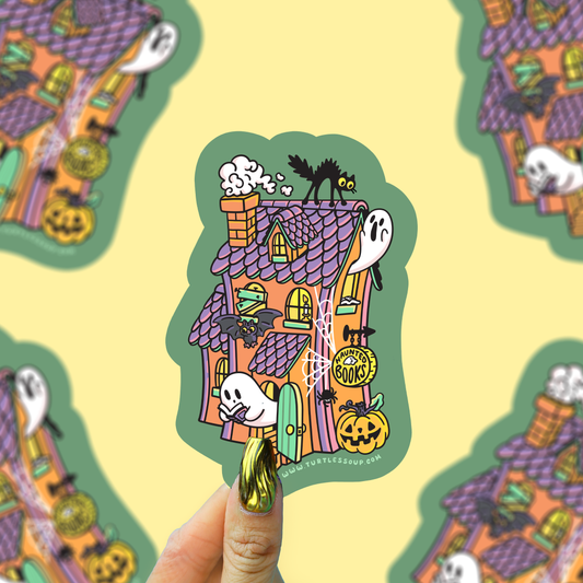 A hand with gold nail polish holds a die-cut sticker featuring a whimsical haunted house illustration with ghosts, pumpkins, a black cat, and a spiderweb on a pale yellow background. The sticker is shown in front of a repeating patterned backdrop.