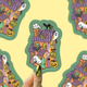 A hand with gold nail polish holds a die-cut sticker featuring a whimsical haunted house illustration with ghosts, pumpkins, a black cat, and a spiderweb on a pale yellow background. The sticker is shown in front of a repeating patterned backdrop.