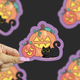 A hand with pink nails holds a die-cut sticker featuring a cartoon jack-o'-lantern, a smaller pumpkin, and a black cat on a dark background with blurred duplicate sticker images.