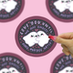 A circular sticker featuring a cute cartoon ghost illustration with the text 'LOVE YOU UNTIL WE ARE GHOSTS AND MAYBE AFTER?' is shown. A hand with red glittery nail polish is touching the sticker.