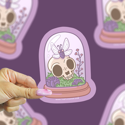 A hand with pink manicured nails holds a die-cut sticker featuring a cartoon skull under a glass dome with a butterfly and purple flowers, set against a purple background with blurred duplicate sticker images.