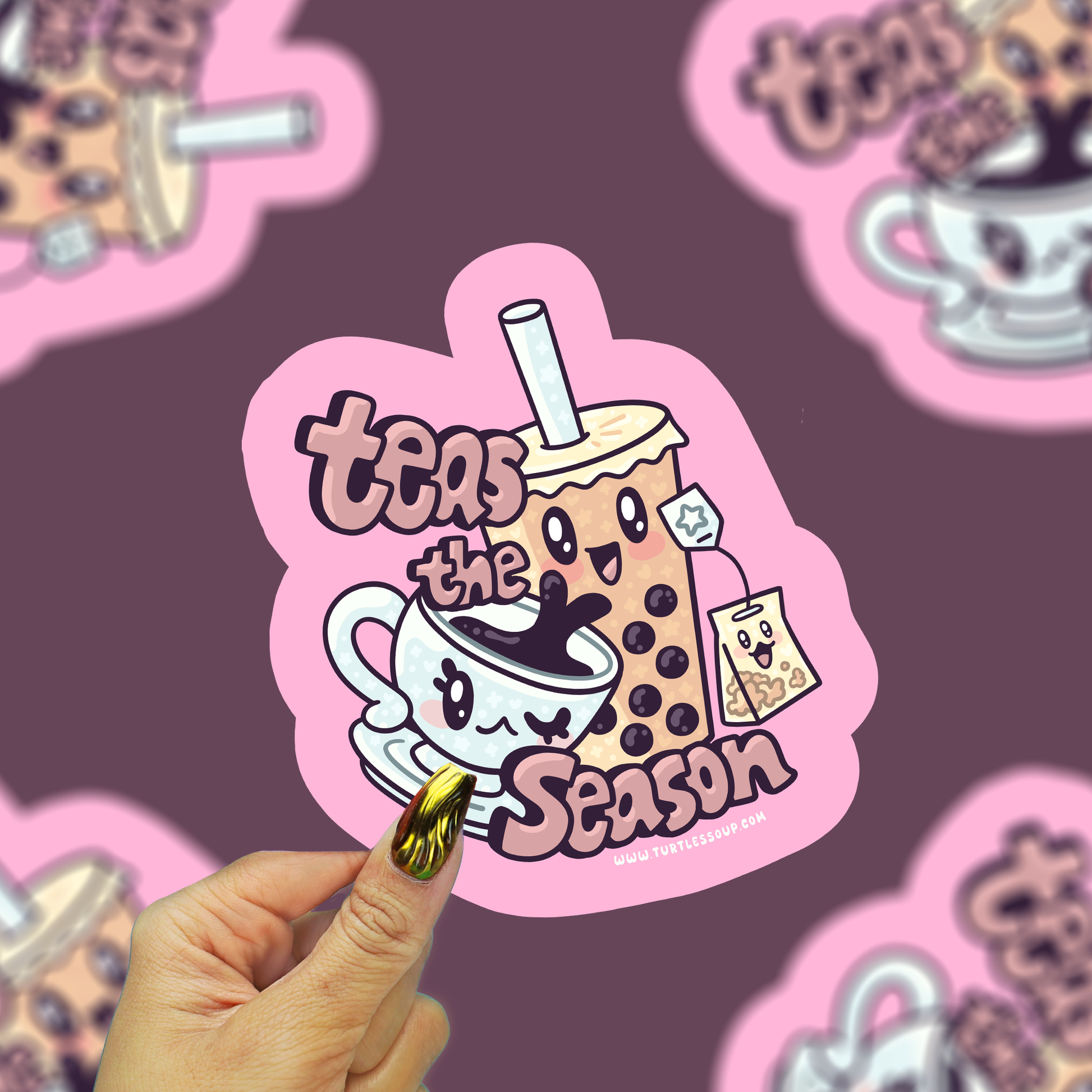 A hand with gold nail polish holds a pink die-cut sticker featuring a cute cartoon boba drink character and the text 'teas the season'. The background is a dark purple surface with several blurred copies of the same sticker.