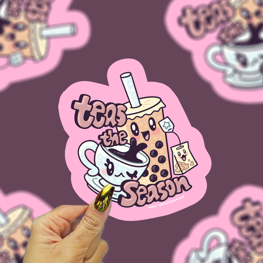 A hand with gold nail polish holds a pink die-cut sticker featuring a cute cartoon boba drink character and the text 'teas the season'. The background is a dark purple surface with several blurred copies of the same sticker.