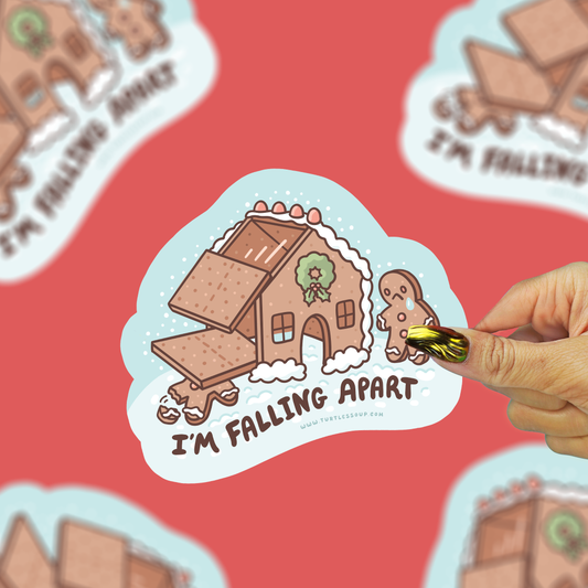 A hand with a gold ring holds a die-cut sticker featuring a gingerbread house and gingerbread man with the text 'I'M FALLING APART'. The sticker is centered against a red background with blurred duplicate sticker images.