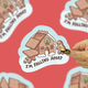 A hand with a gold ring holds a die-cut sticker featuring a gingerbread house and gingerbread man with the text 'I'M FALLING APART'. The sticker is centered against a red background with blurred duplicate sticker images.