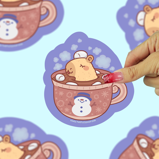 A hand with pink nail polish peels a die-cut sticker from a light blue background patterned with the same sticker design. The sticker features a cute hamster sleeping in a hot cocoa cup with a snowman on the cup, set against a light blue background.